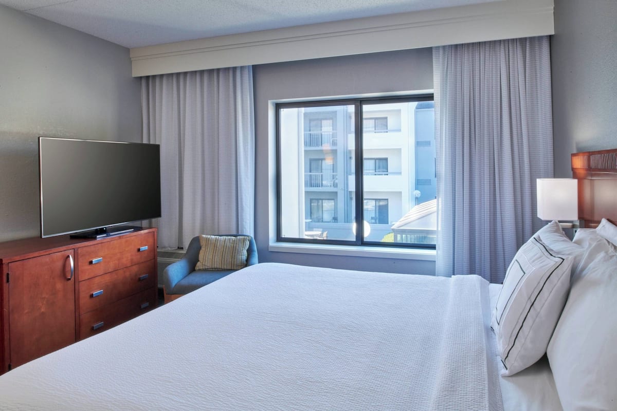 A hotel room with a large bed, a flat-screen TV on a wooden dresser, a window with curtains, and a view of a building outside.