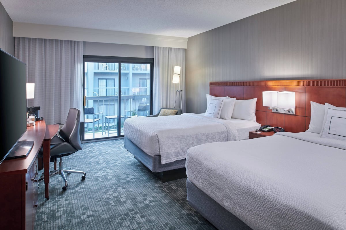 A hotel room with two beds, a desk, a chair, a TV, and a balcony with a view.