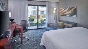 Quarto de hotel com uma cama, uma escrivaninha com computador, uma cadeira, um sofá, um abajur e um quadro na parede.