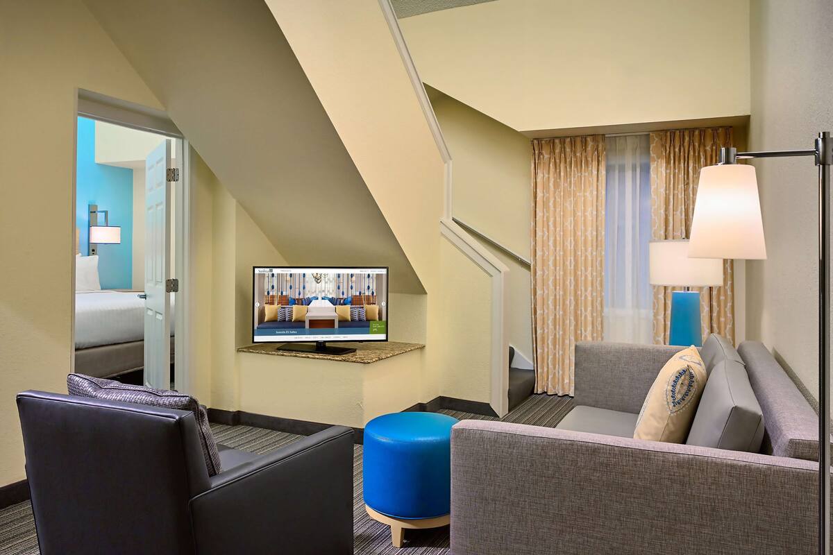 Une chambre d’hôtel équipée d’un canapé, d’un pouf et d’un téléviseur affichant un plan de la chambre.