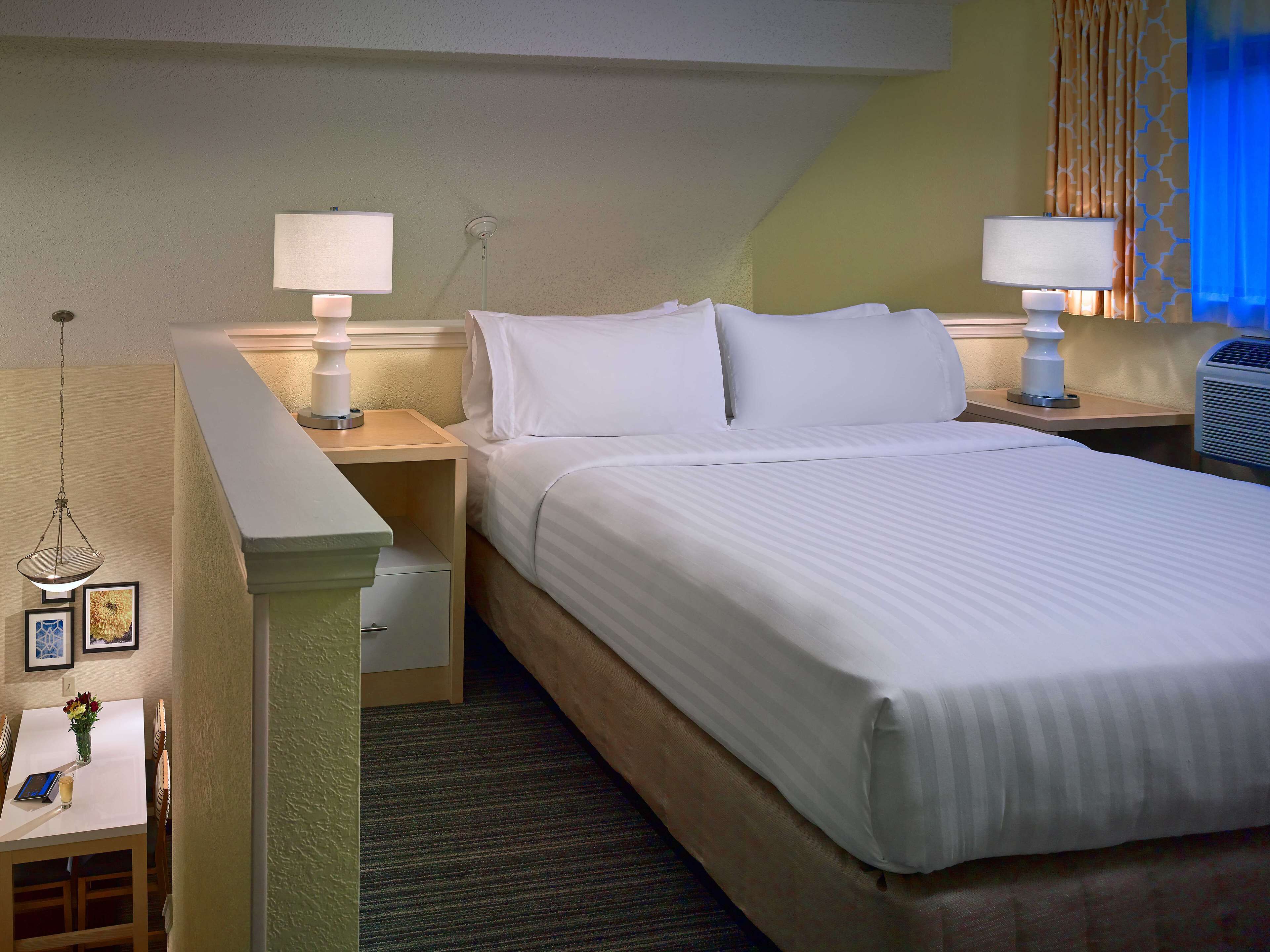 A hotel room with a neatly made bed, two bedside lamps, a small table with a vase of flowers, and a picture frame.