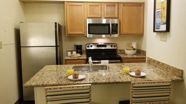 A compact kitchen with a refrigerator, microwave, stove, and granite countertop. There are two chairs and a small island with a sink. - Sonesta Simply Suites Memphis East Poplar Pike ​ (Memphis)