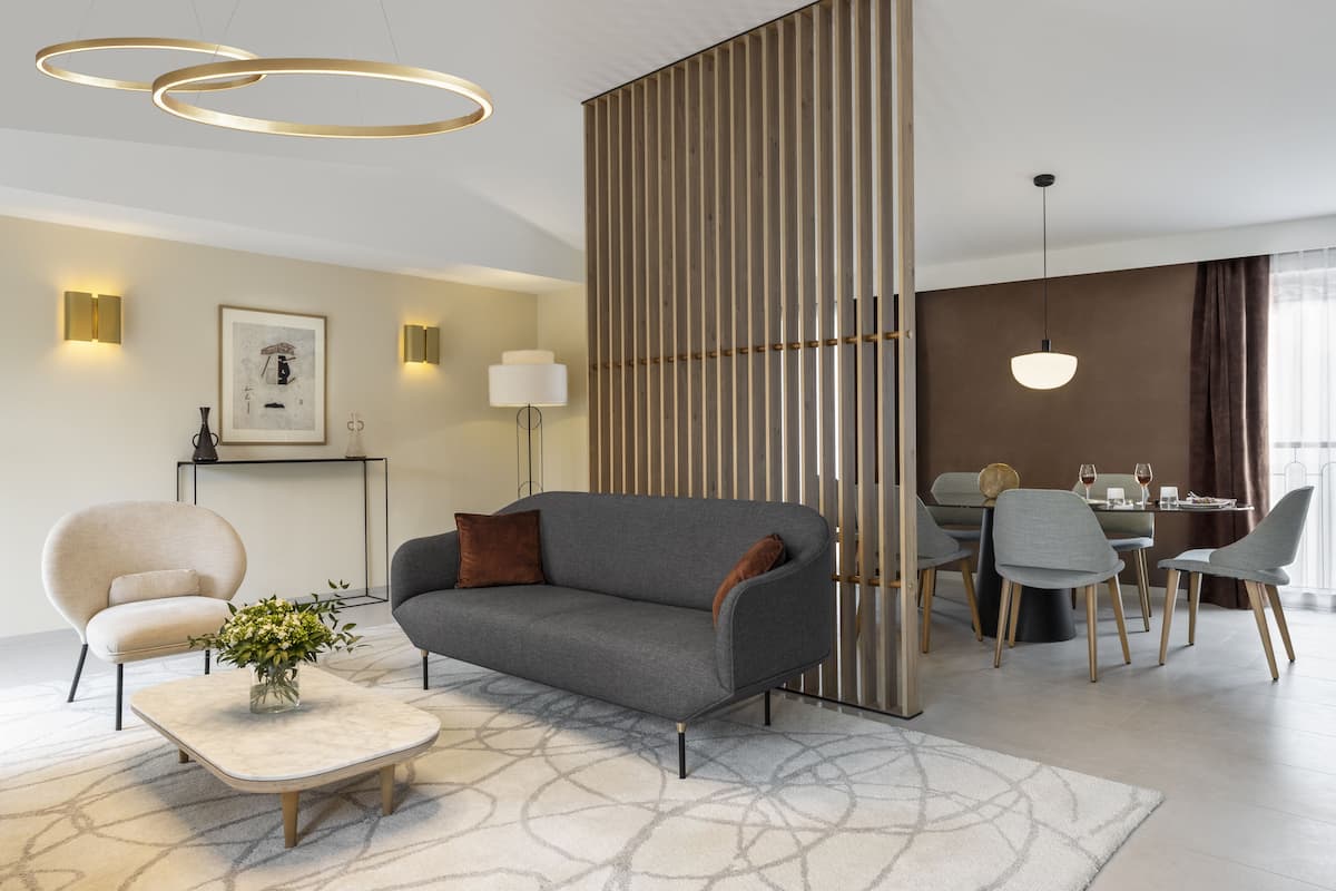 A modern living room with a grey sofa, a beige armchair, and a coffee table with a vase of flowers. There is a dining area with a table and chairs, and a decorative wall panel.