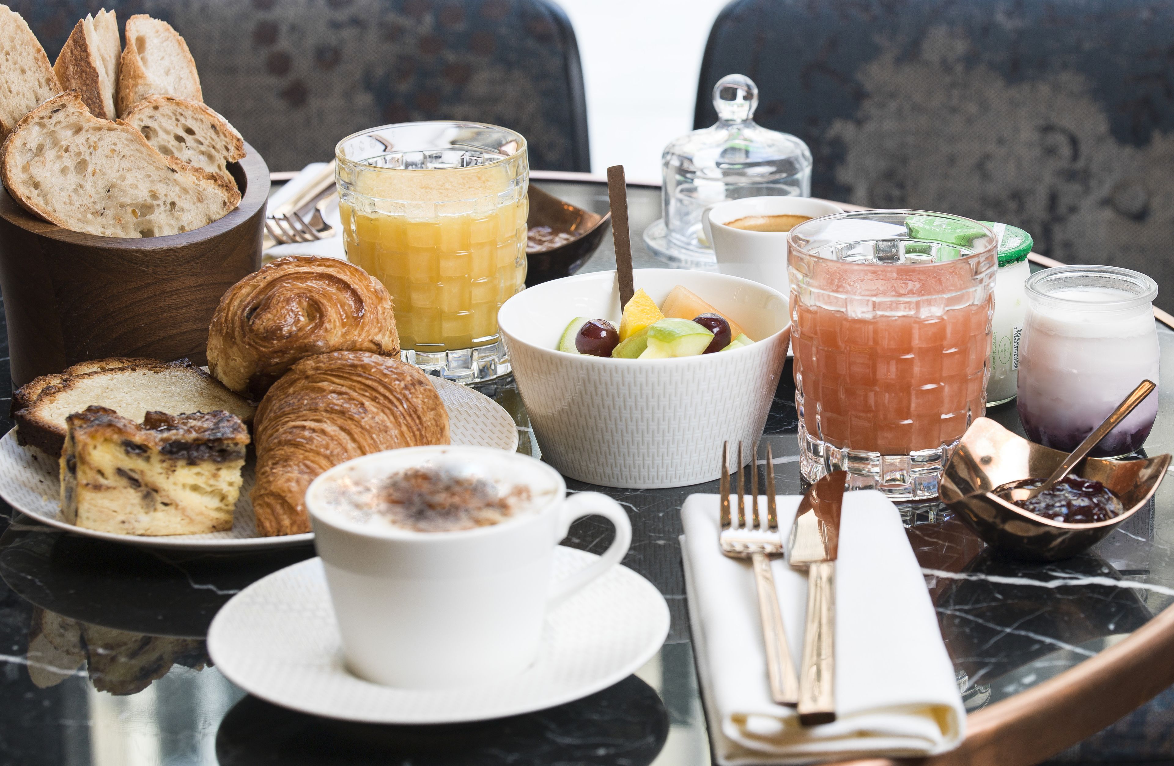 A breakfast table with croissants, bread, a fruit bowl, a glass of orange juice, a glass of iced tea, a cup of coffee, and a glass of milk.