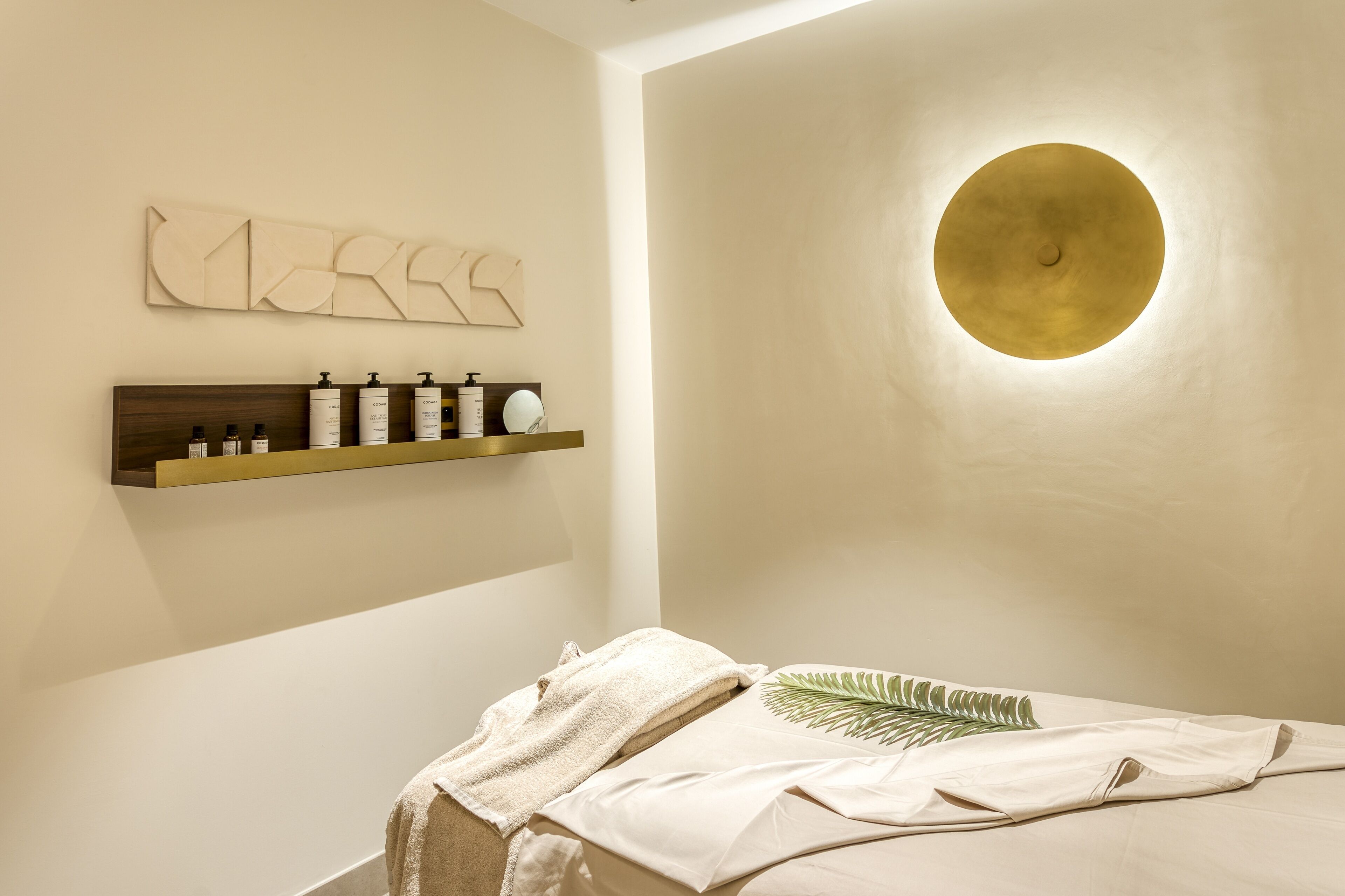 A spa room with a wooden shelf holding toiletries, a circular wall light, and a bed with a towel and leaf-patterned pillow.