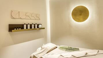 A spa room with a wooden shelf holding toiletries, a circular wall light, and a bed with a towel and leaf-patterned pillow.