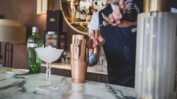 Un barman prépare un cocktail dans un bar doté d’un comptoir en marbre, d’une bouteille verte et d’un shaker à cocktail doré.