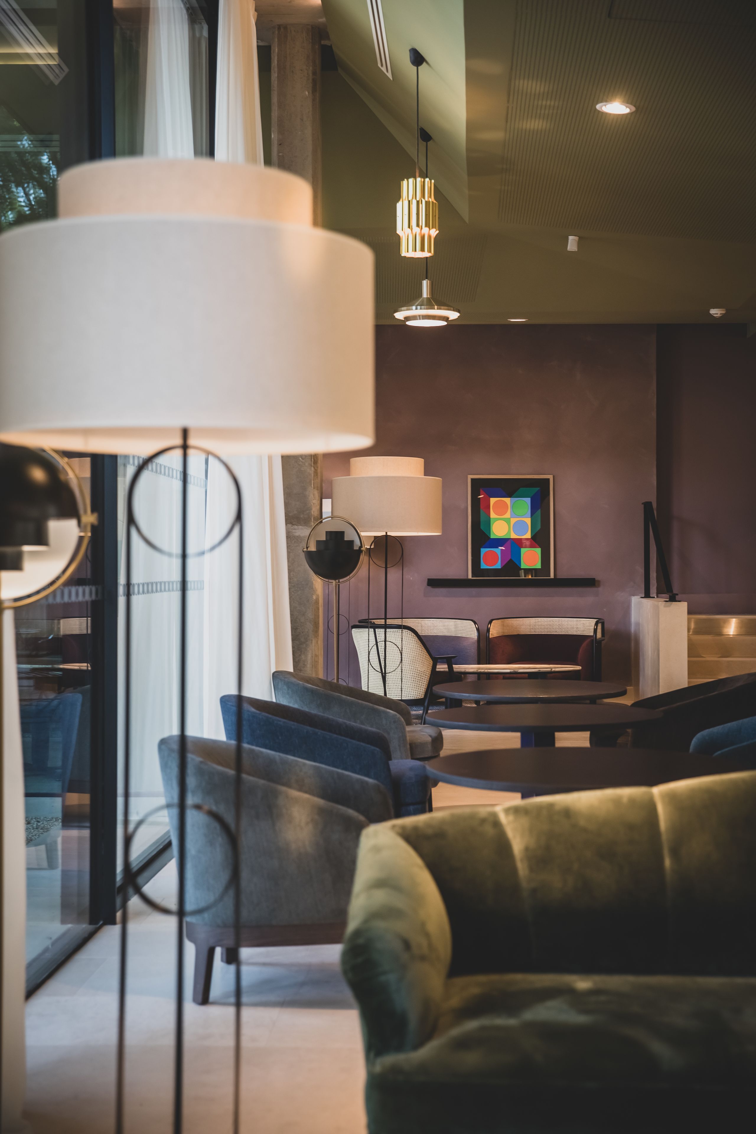 A modern lounge area with a sofa, chairs, and a table. There are two pendant lights and a wall-mounted lamp. A colorful abstract painting is visible on the wall.