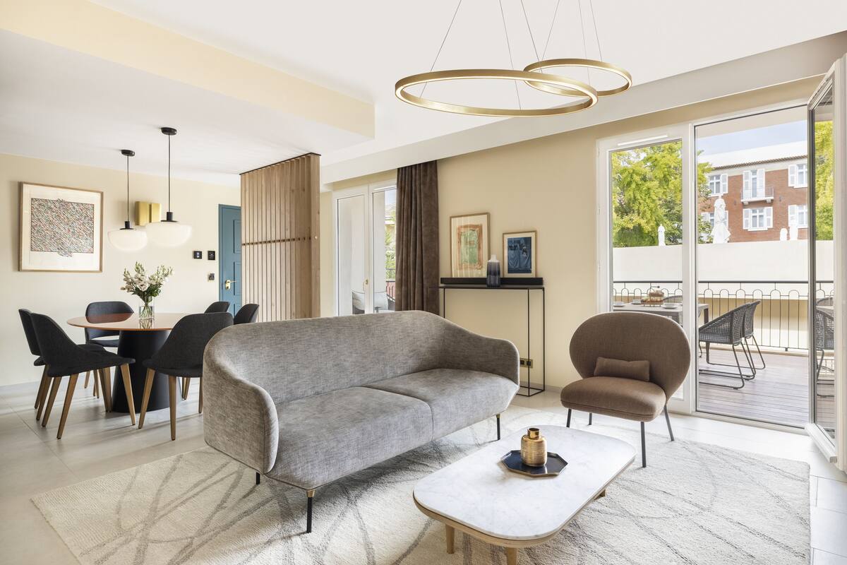 A modern living room with a grey sofa, a round coffee table, and a dining area with a table and chairs. There is a large window with a view of a balcony and buildings outside.