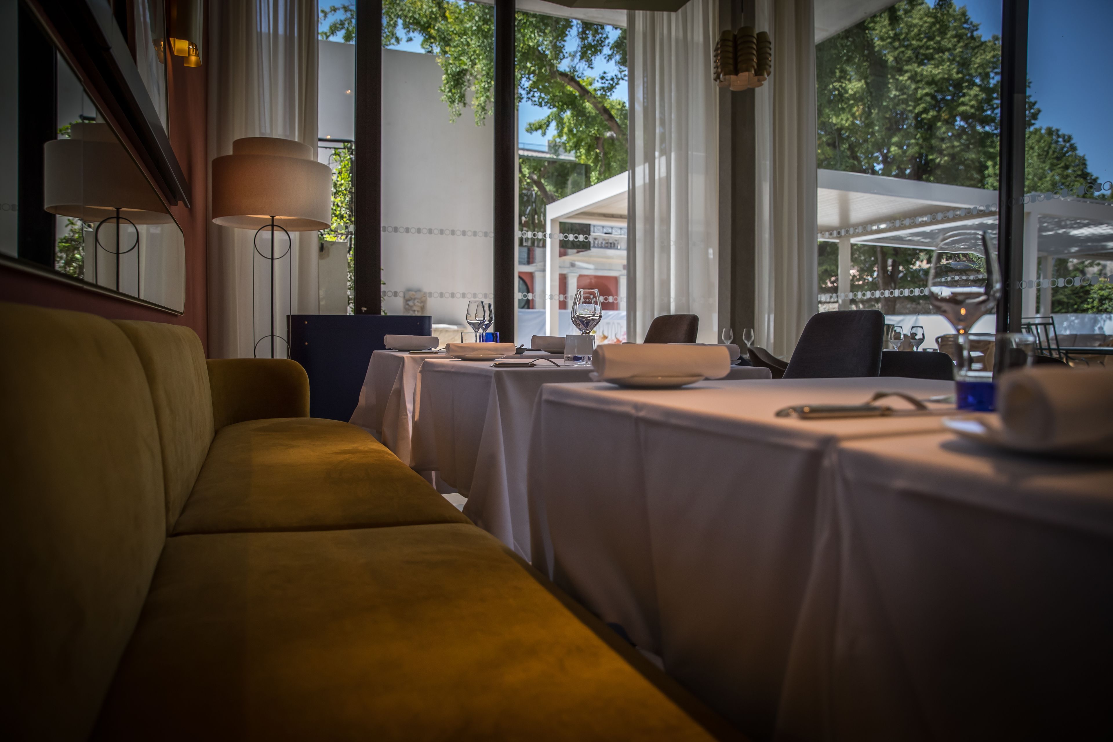 A restaurant with a yellow sofa, tables set with white tablecloths, and large windows offering a view of the outdoors.