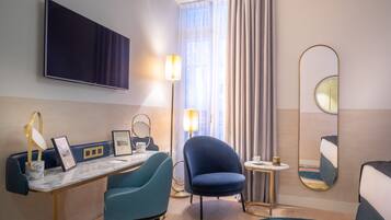 A modern hotel room with a flat-screen TV, a desk with a lamp, a comfortable chair, a small round table, and a large mirror.
