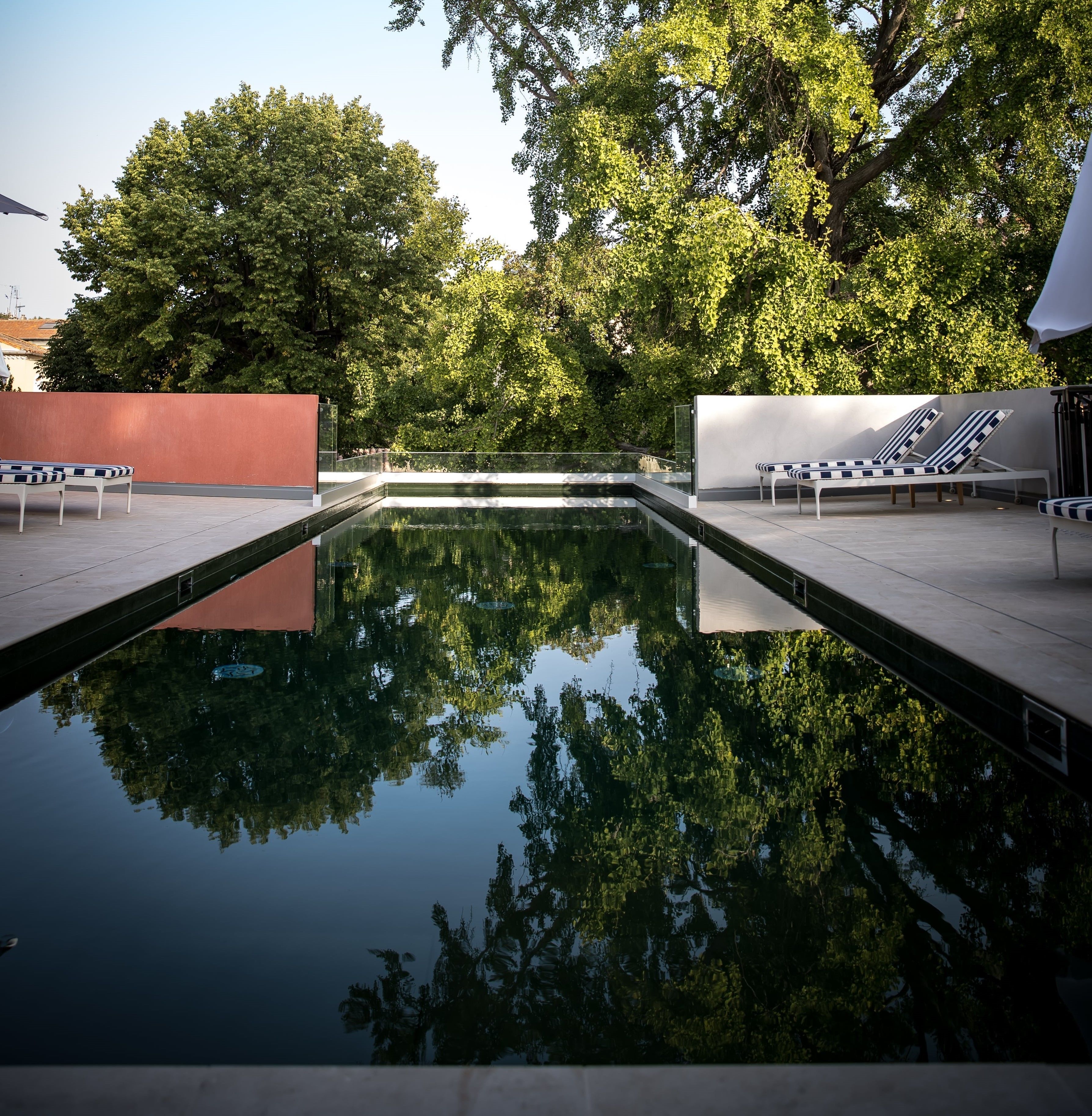 A rectangular pool with clear reflections of surrounding trees, two lounge chairs, and a red wall.