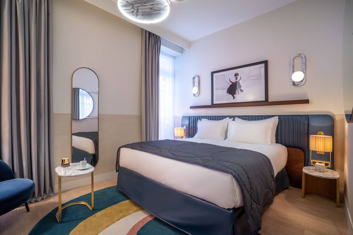 A modern hotel room with a large bed, a round mirror, a framed picture on the wall, and a bedside table with a lamp.