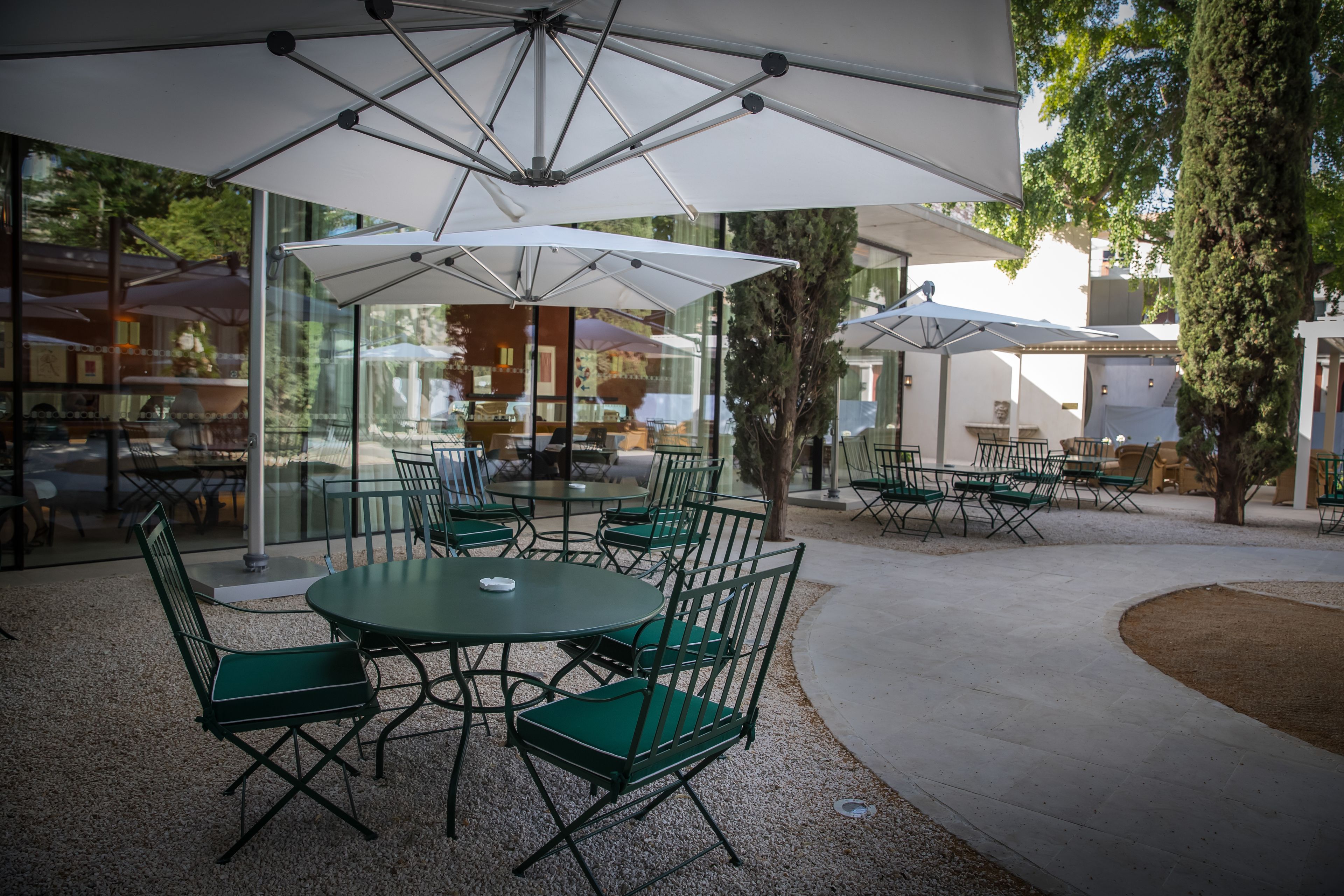 An outdoor seating area with round tables and green chairs, covered by white umbrellas, and surrounded by glass walls.