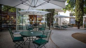 An outdoor seating area with round tables and green chairs, covered by white umbrellas, and surrounded by glass walls.