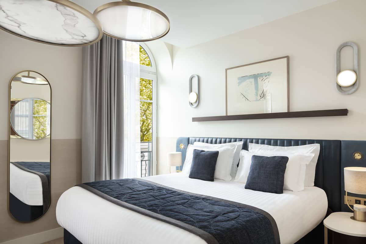 A modern hotel room with a large bed, a mirror, a bedside table with a lamp, and a window with curtains.