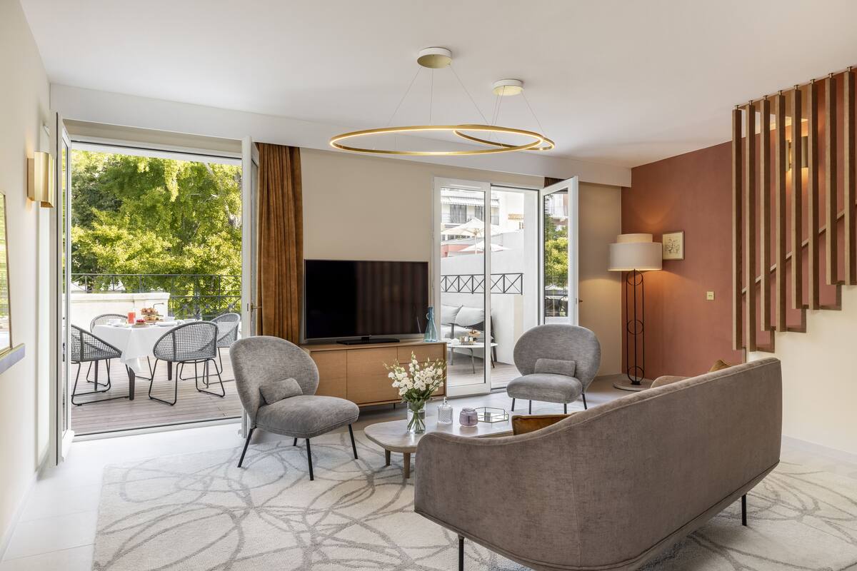 A modern living room with a sofa, armchairs, a coffee table, and a TV. There is a balcony with outdoor furniture.