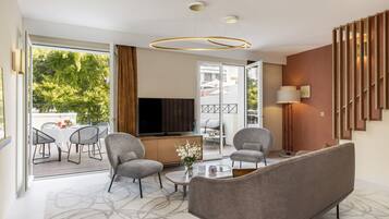 Ein modernes Wohnzimmer mit Sofa, Sesseln, Couchtisch und Fernseher. Ein Balkon mit Gartenmöbeln ist vorhanden.