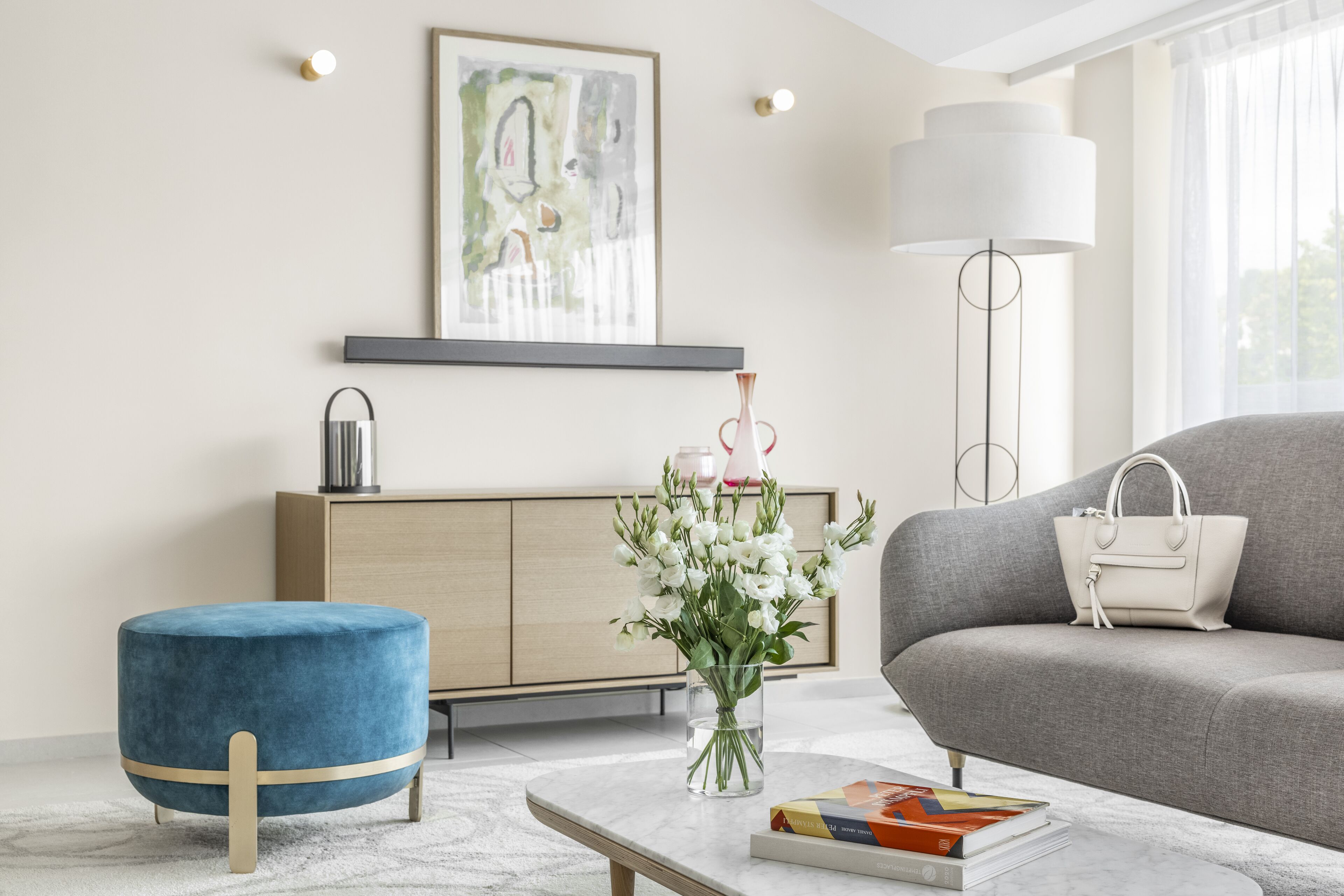 Una sala moderna con un sofá, un reposapiés azul, un jarrón de flores, una lámpara y una estantería.