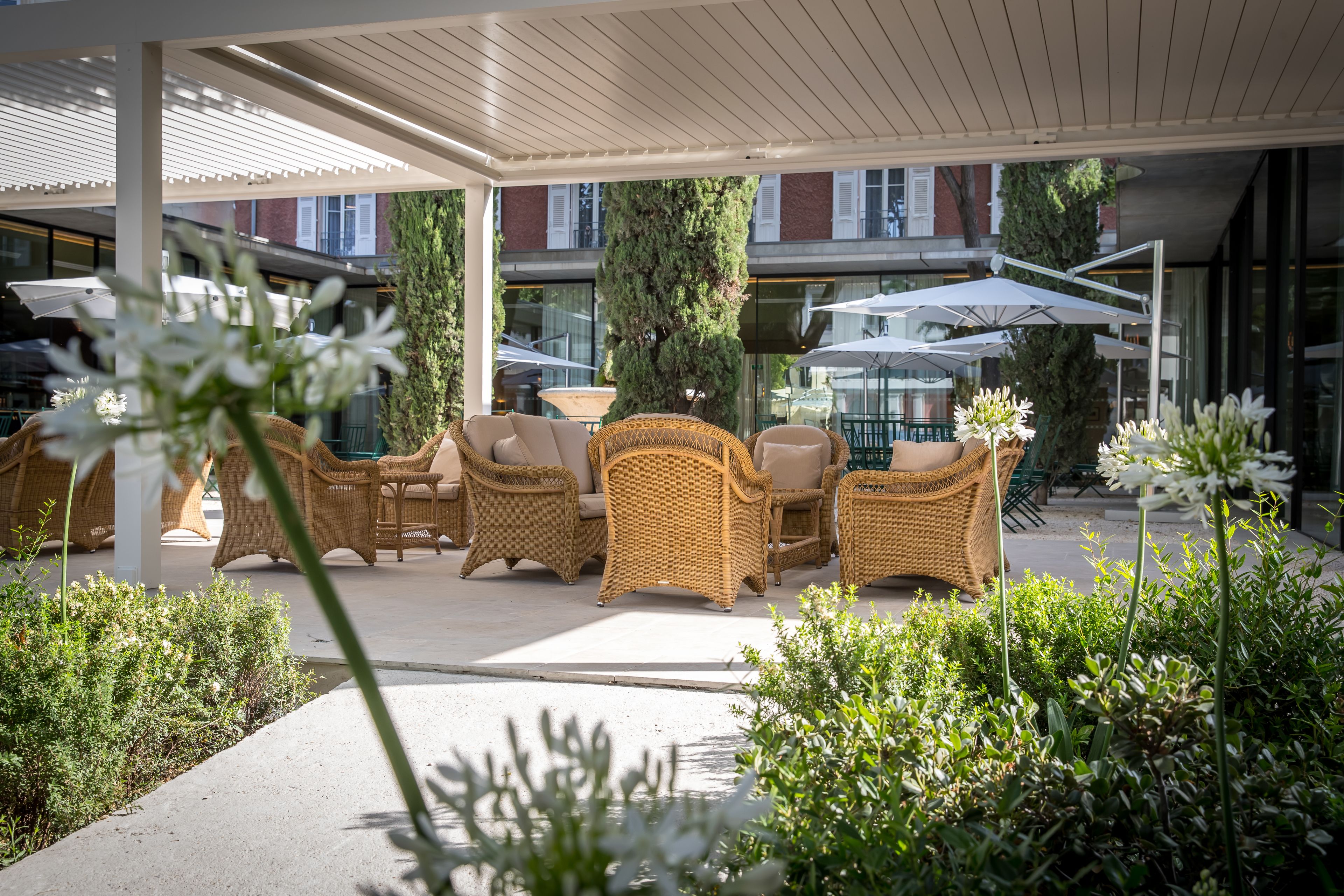 Un espace extérieur aménagé avec des meubles en osier et des parasols, entouré de verdure et bordé d’une allée couverte.