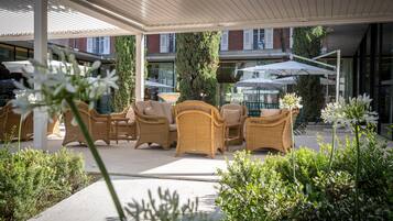 Un espace extérieur aménagé avec des meubles en osier et des parasols, entouré de verdure et bordé d’une allée couverte.