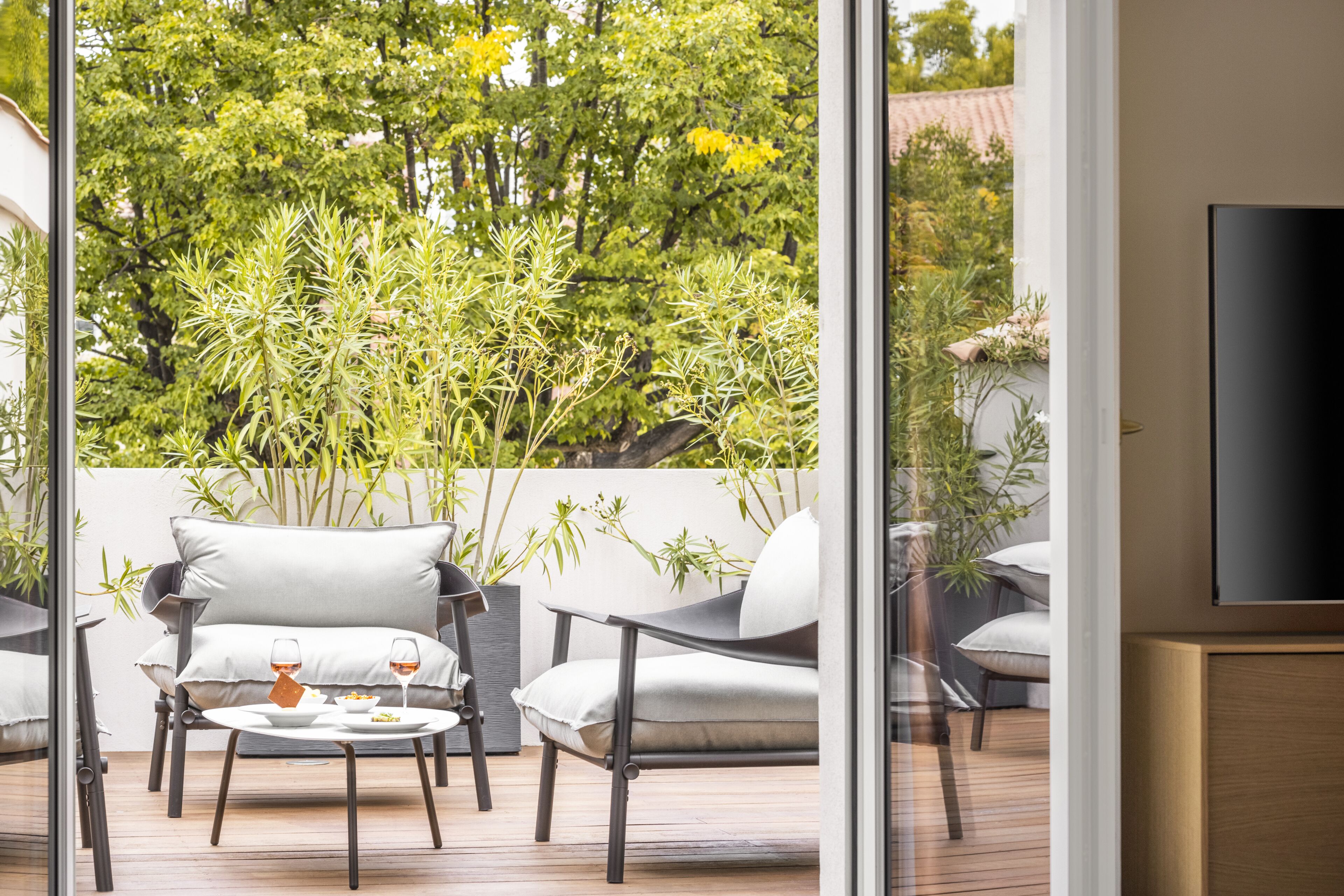 A modern outdoor seating area with two chairs and a table, a flat-screen TV mounted on the wall, and lush greenery in the background.