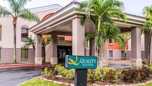 A hotel entrance with a sign reading "Quality Suites," featuring a covered entrance, palm trees, and a well-maintained lawn. - Quality Suites Fort Myers I-75 (Fort Myers)