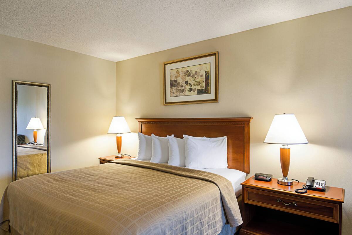 A hotel room with a bed, two bedside lamps, a bedside table with a phone, a mirror, and a framed picture on the wall.