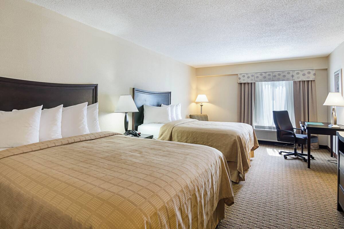 A hotel room with two beds, a desk, a chair, and a lamp.