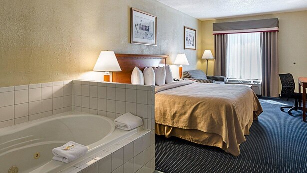 A hotel room with a large bed, a sitting area with a chair, a desk, a window with curtains, and a bathtub with a towel. - Quality Inn Troutville - Roanoke North (Troutville)