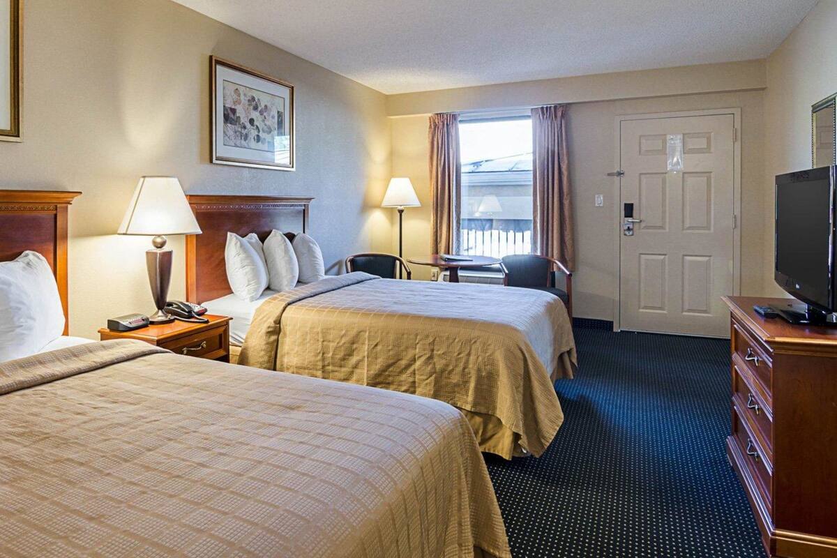 A hotel room with two beds, a desk, a chair, a TV, and a door.