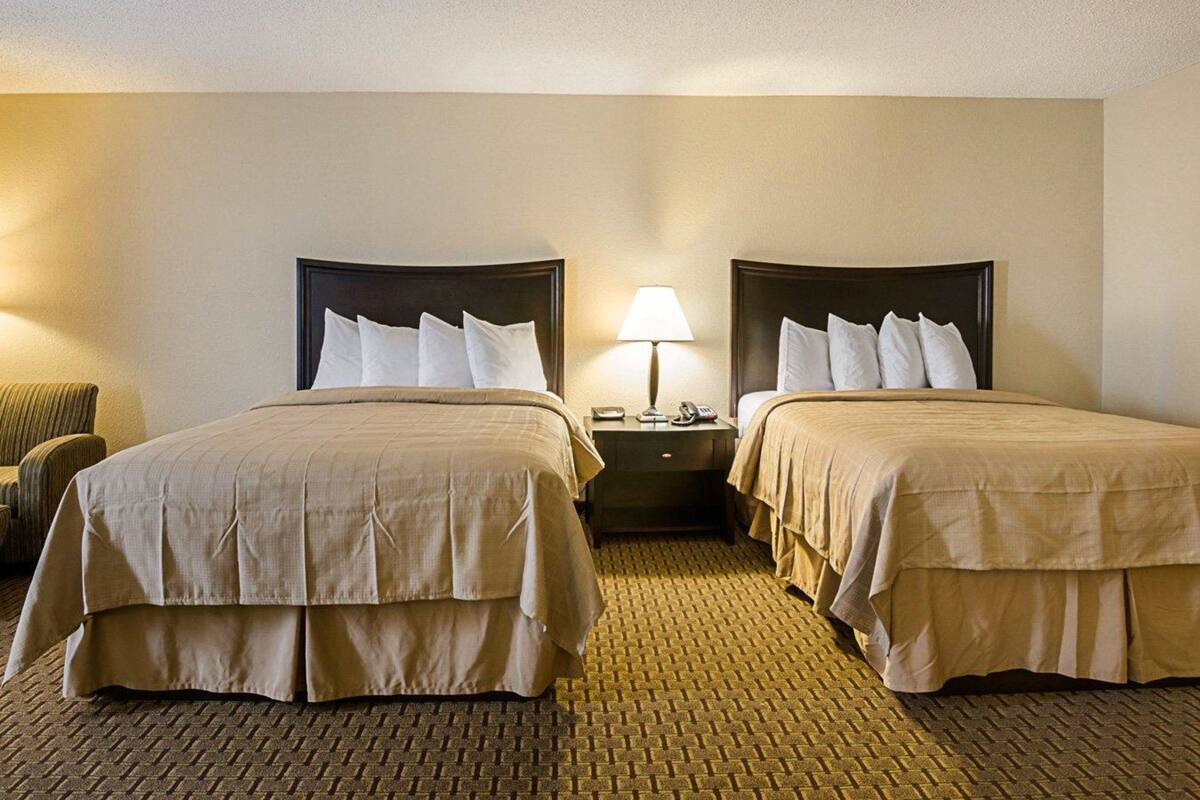 A hotel room with two beds, a nightstand with a lamp, and a chair.