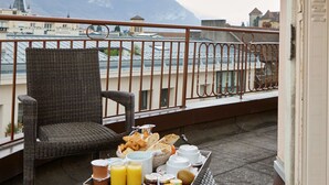 Un balcón con mobiliario de mimbre, una bandeja con alimentos para el desayuno y vistas a edificios y montañas al fondo.