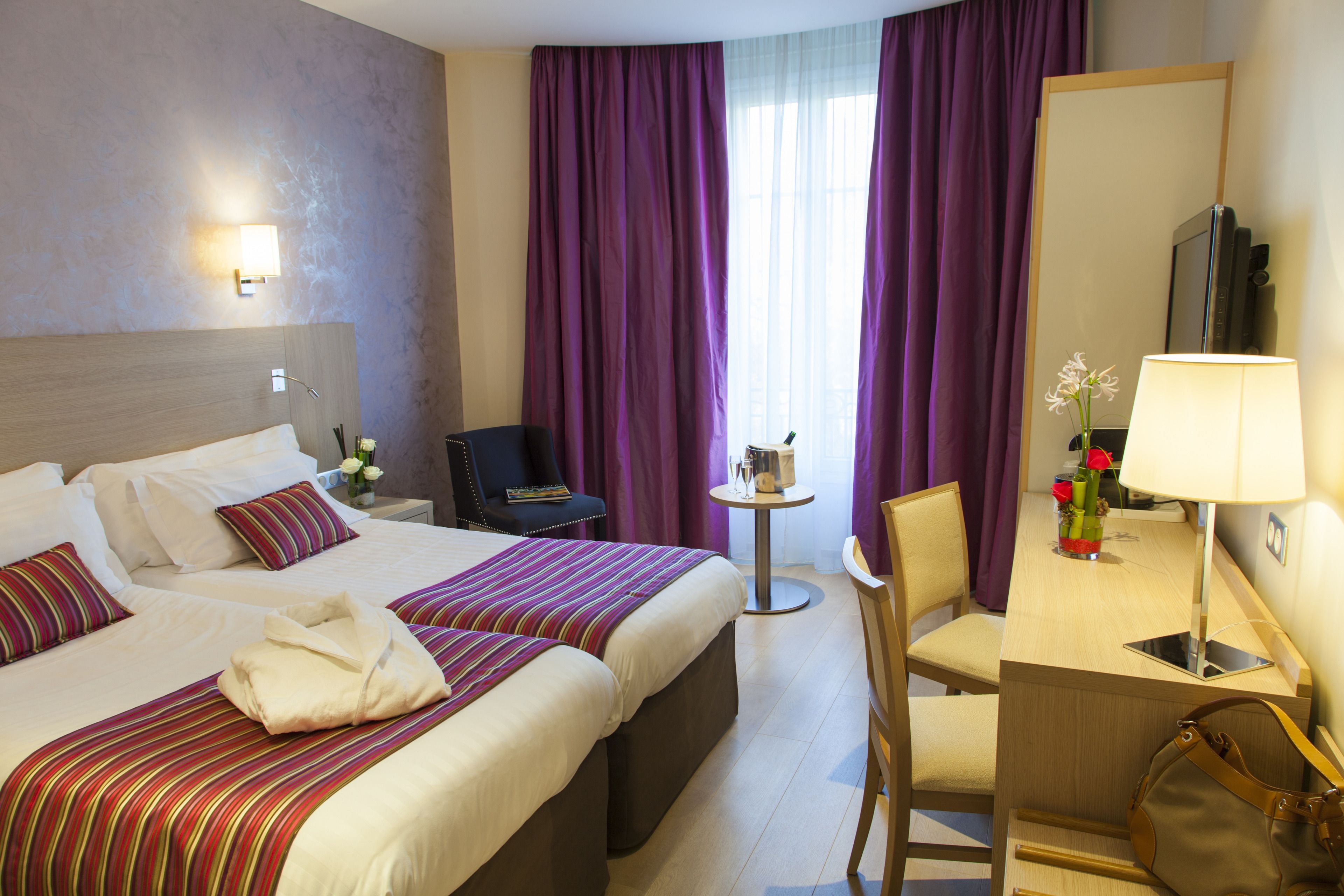 A hotel room with two beds, a desk, a chair, a mirror, and a window with purple curtains.