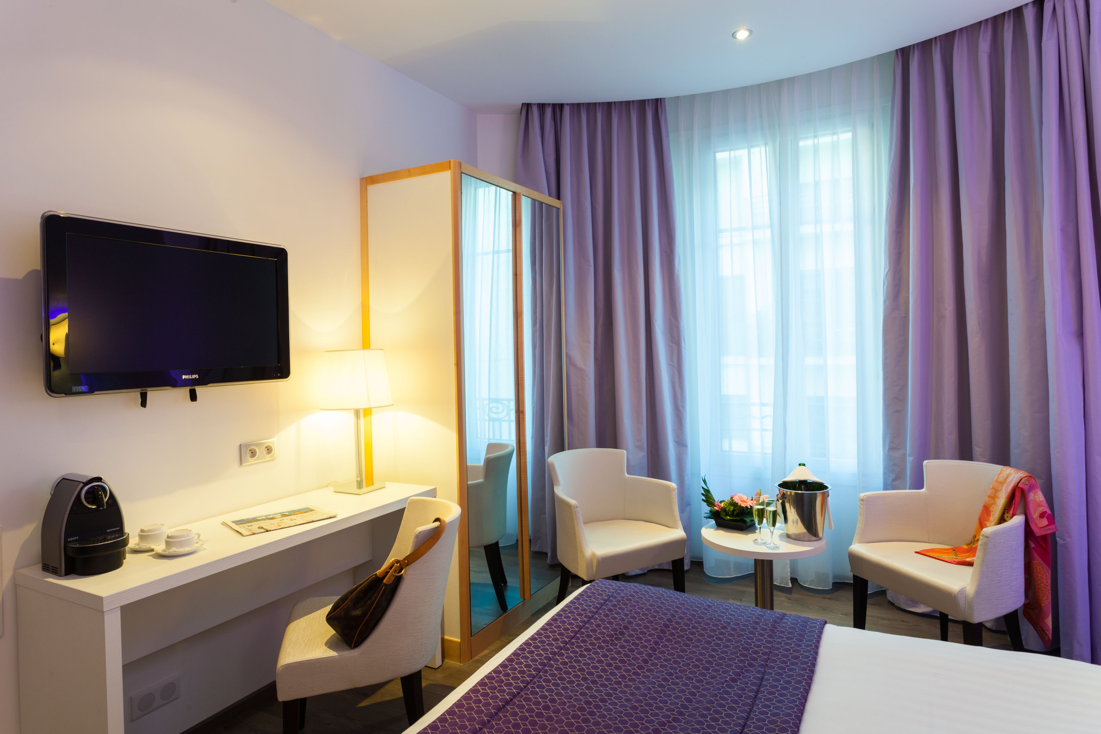 A hotel room with a bed, a desk with a computer, a chair, a TV, and a window with curtains.