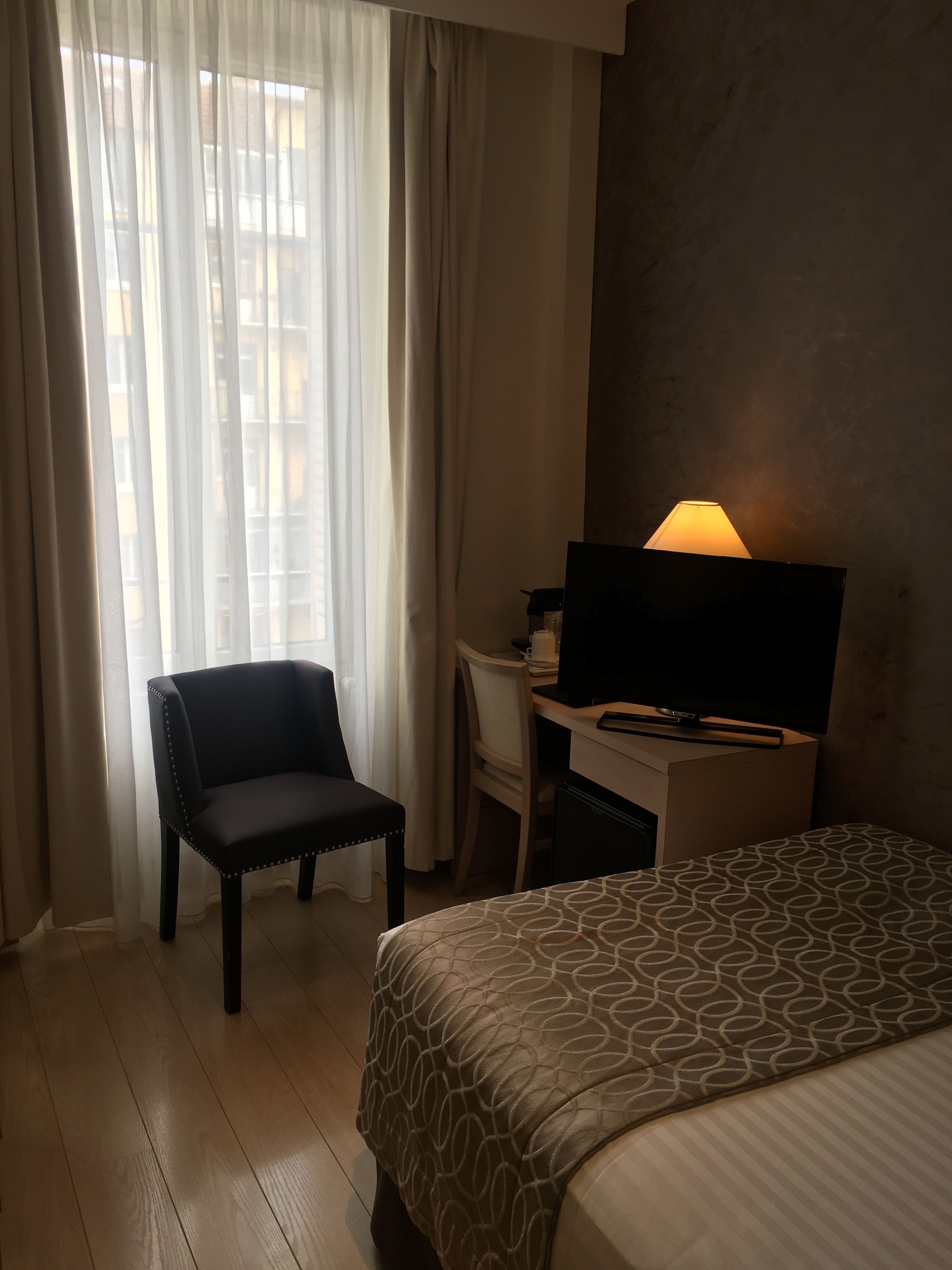 A hotel room with a bed, a desk with a television, a chair, and a window with curtains.