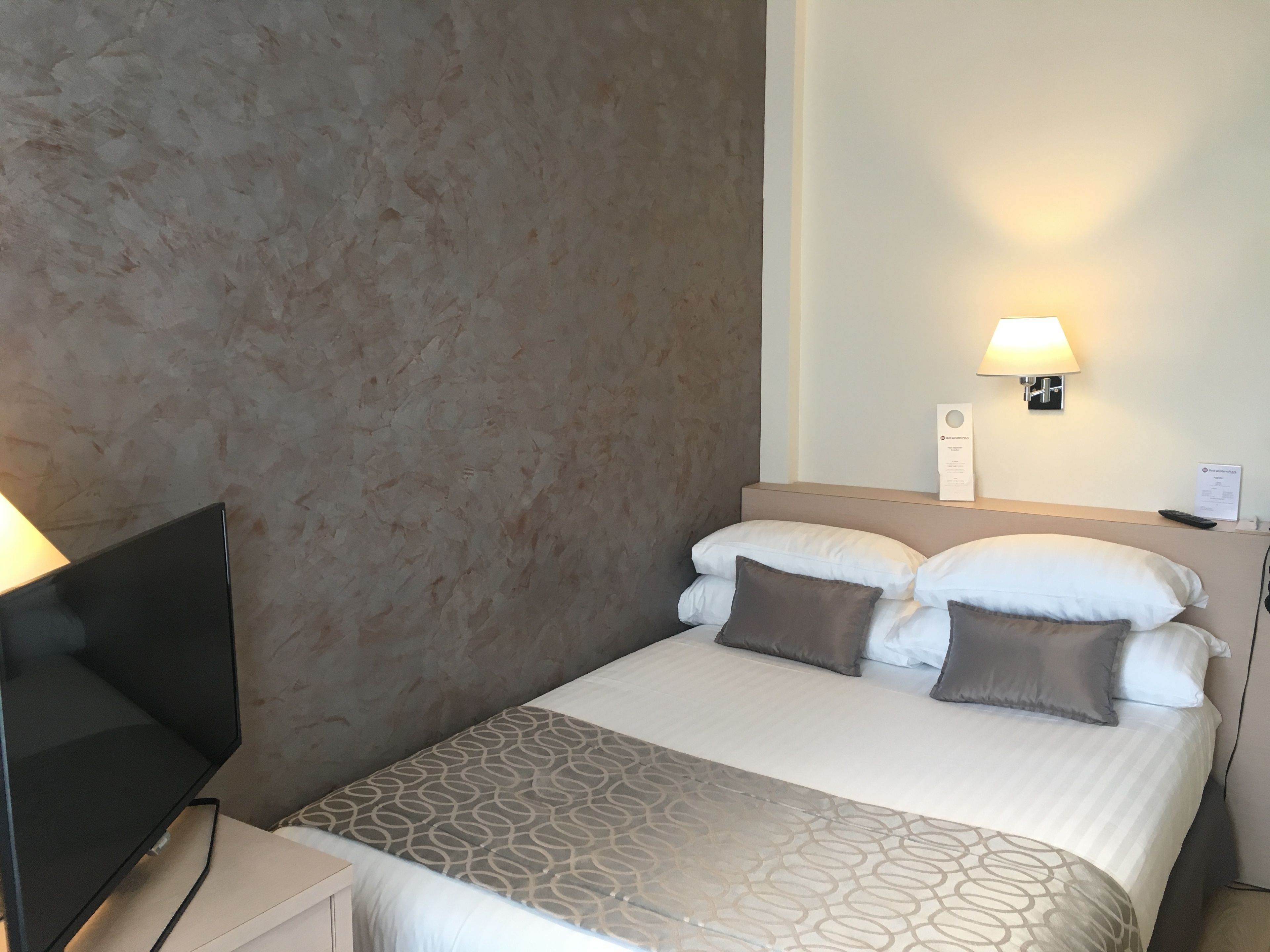 Een hotelkamer met een bed, een televisie, een lamp en een bedieningspaneel aan de muur.
