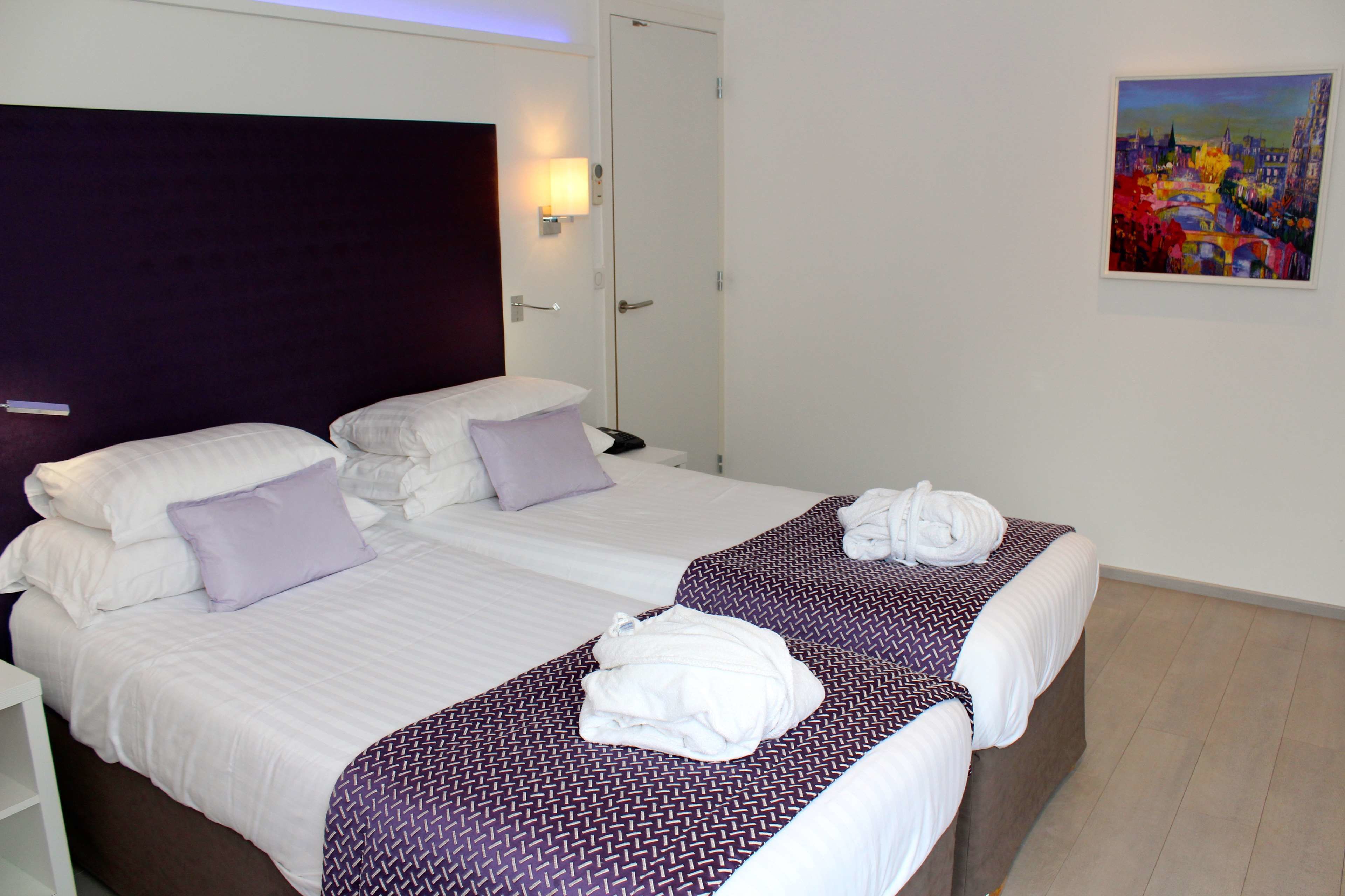 Habitación de hotel con dos camas, un cabecero oscuro, un cuadro en la pared y una puerta con lámpara.