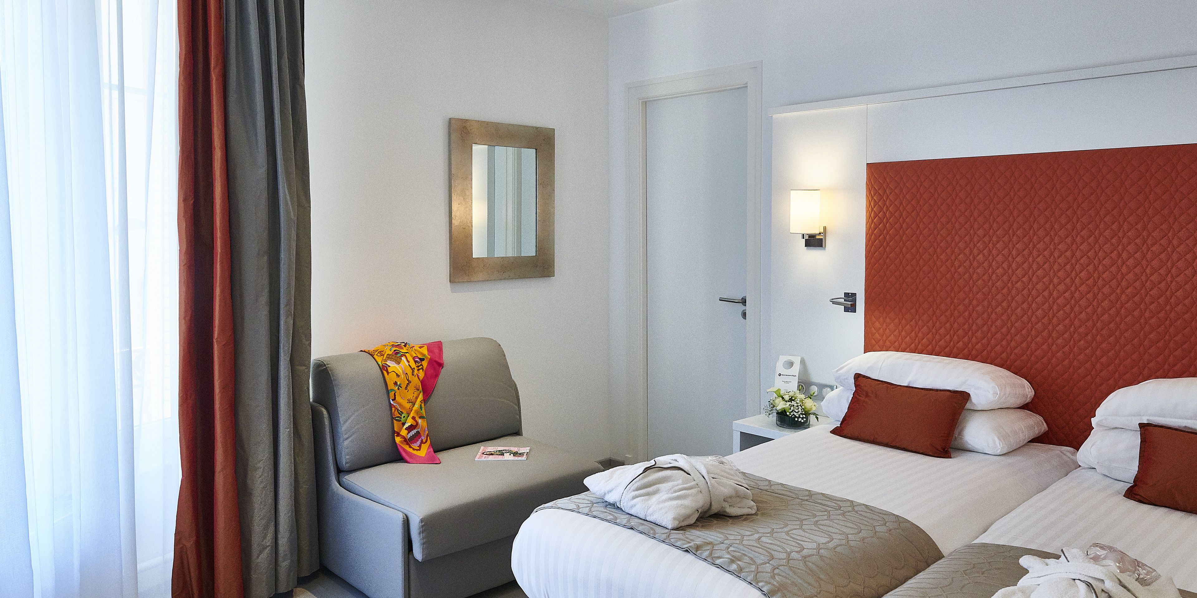 A hotel room with a grey sofa, a bed with white and brown pillows, a small bedside table with a lamp, and a door leading to another room.