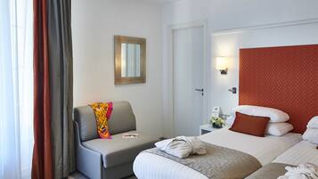 A hotel room with a grey sofa, a bed with white and brown pillows, a small bedside table with a lamp, and a door leading to another room.