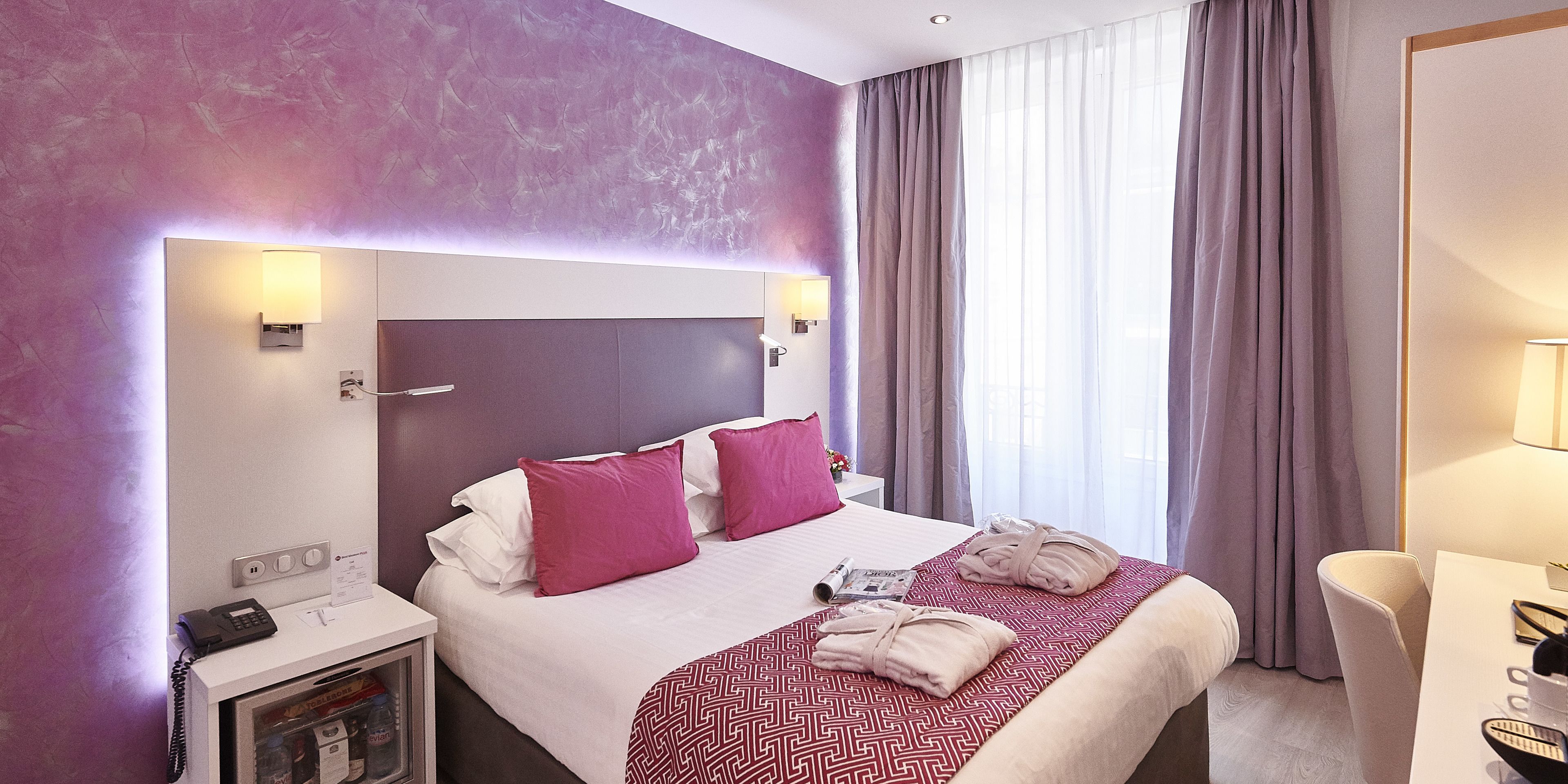 Een hotelkamer met een groot bed, een nachtkastje met telefoon, een minibar en uitzicht buiten door een raam met gordijnen.