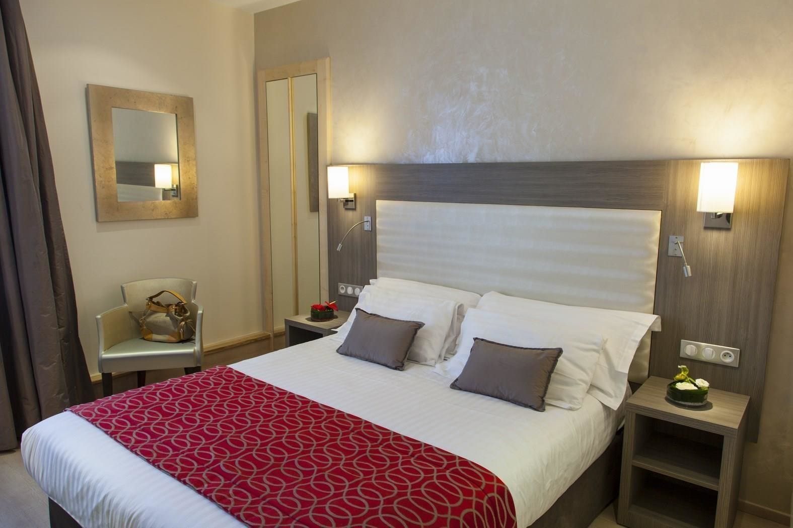 A hotel room with a large bed, two bedside tables, a chair, a mirror, and a nightstand with a lamp.