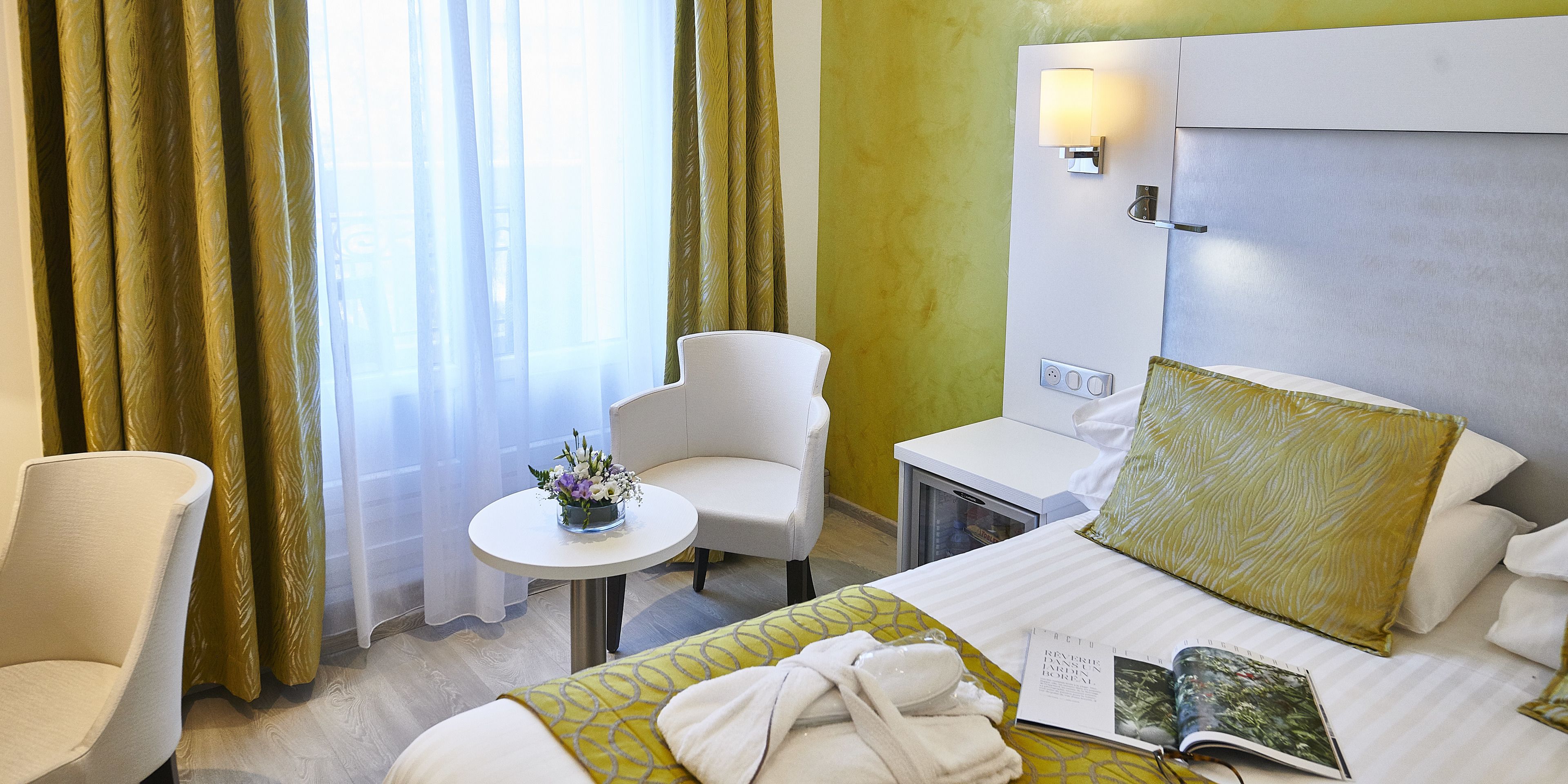 A hotel room with a bed, a bedside table, a chair, a small table with a vase of flowers, and a window with curtains.