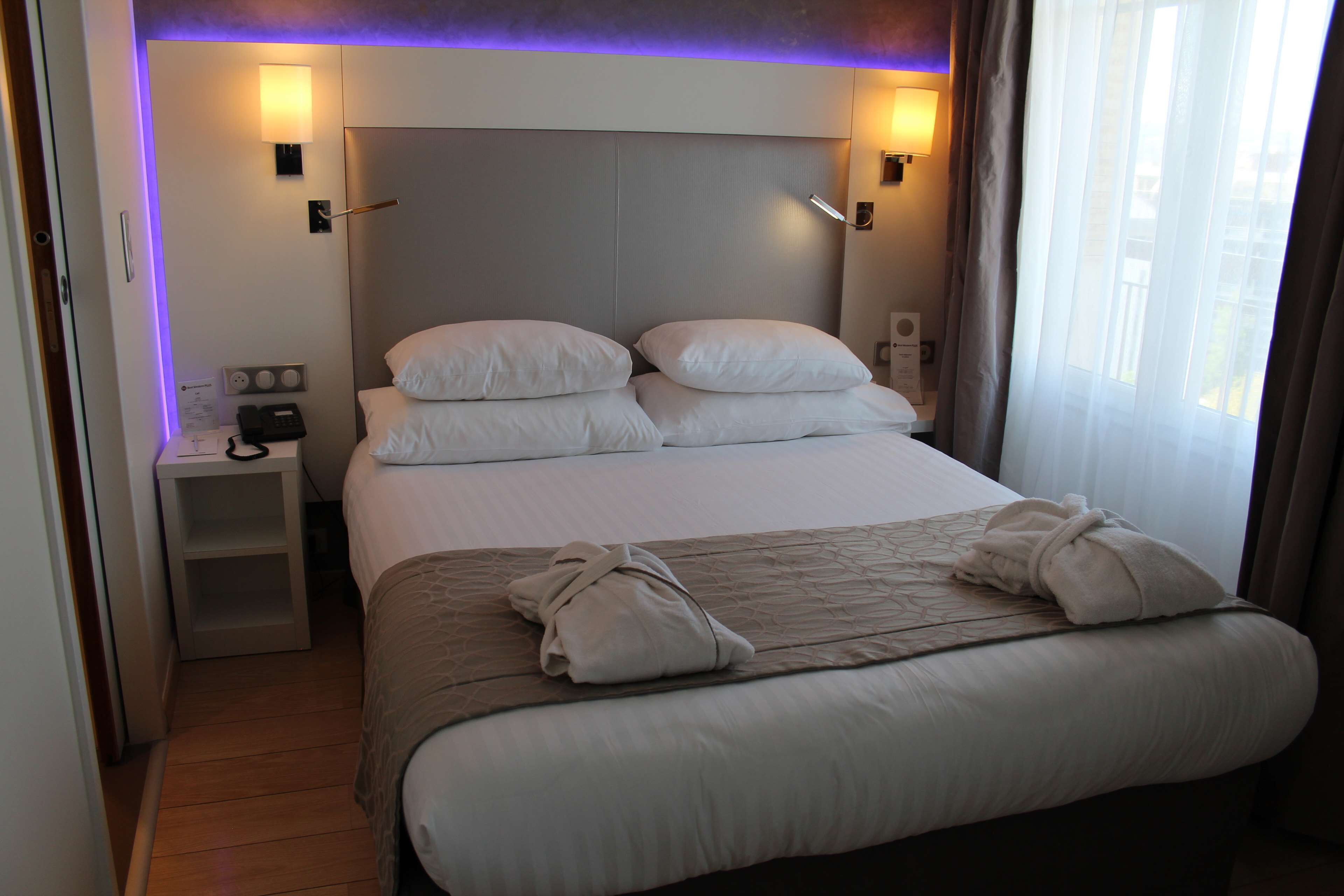A hotel room with a large bed, two bedside tables, a wall-mounted lamp, and a window with curtains.