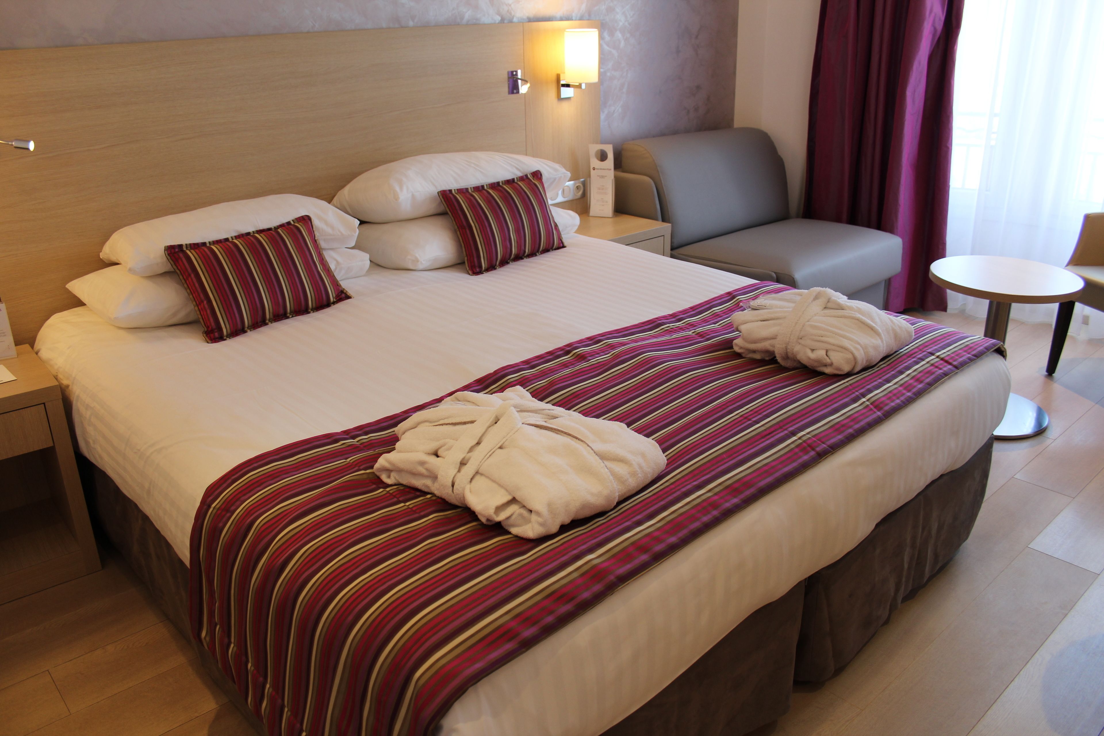 A hotel room with a large bed, two pillows, a bedside table with a lamp, a chair, and a small round table.