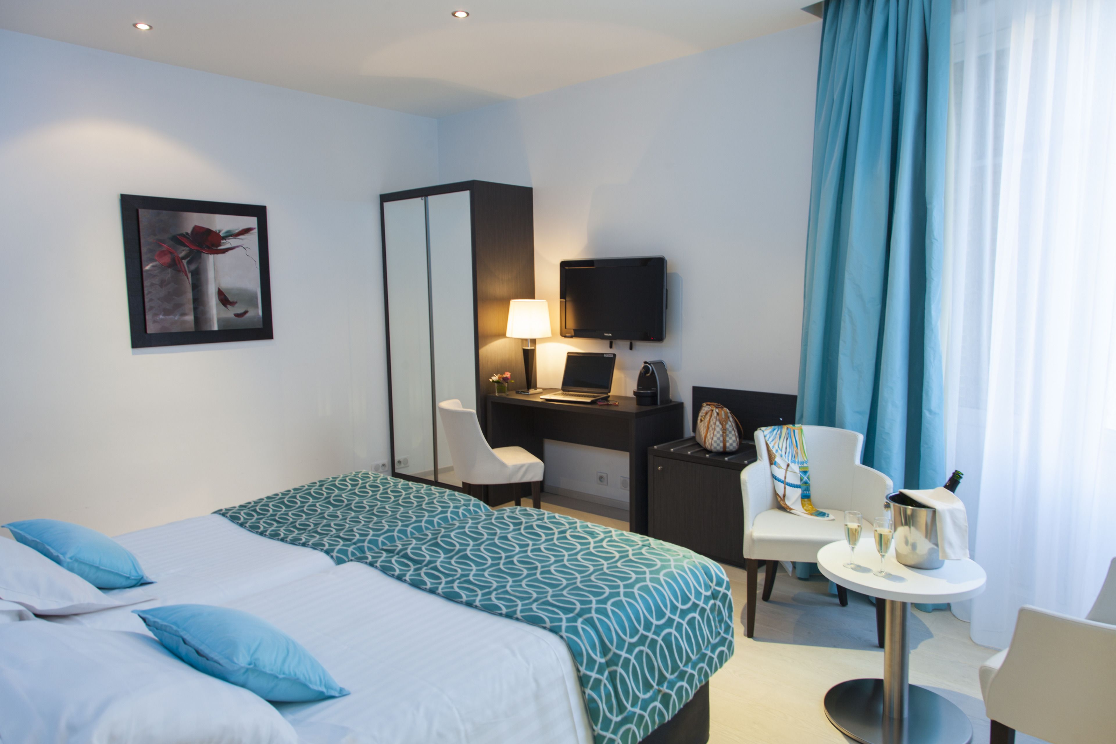 A hotel room with a bed, a desk with a computer, a chair, a small table with a bottle and glasses, and a TV on the wall.