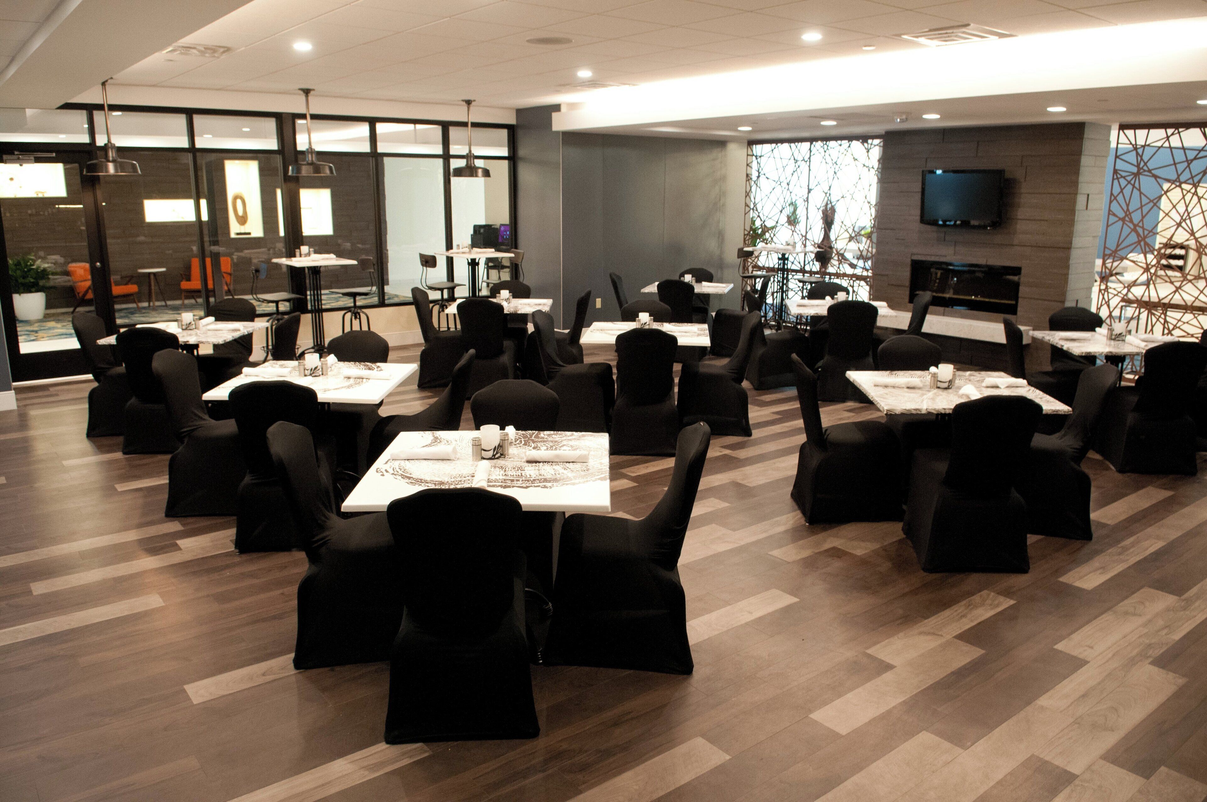 Un espace repas moderne doté de plusieurs tables et chaises, d’un téléviseur à écran plat fixé au mur et offrant une vue sur une fenêtre à motifs.