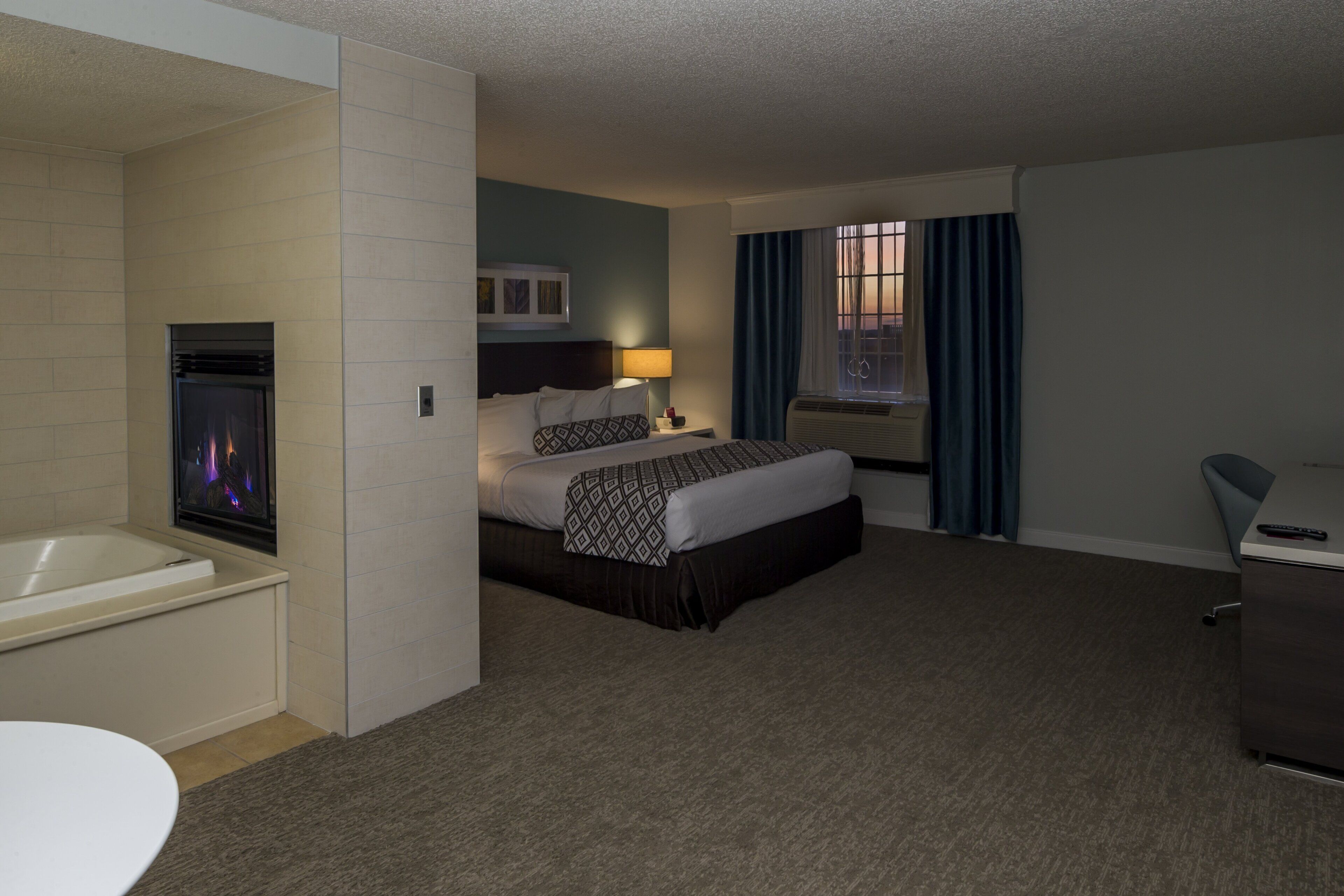 Habitación de hotel con cama, escritorio, silla, baño con bañera y ventana con cortinas.