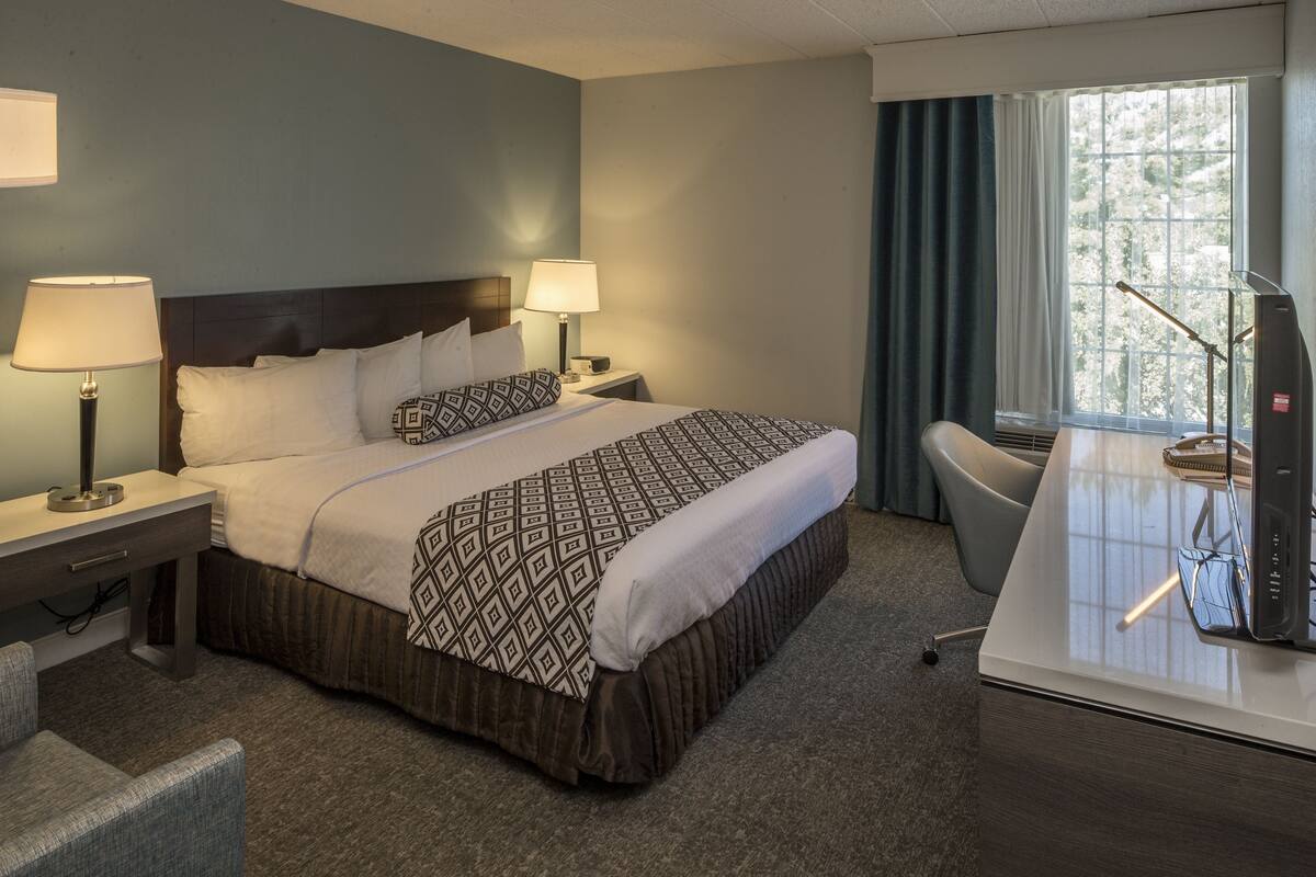 Ein Hotelzimmer mit einem Bett, zwei Nachttischlampen, einem Schreibtisch mit Computer und einem Fenster mit Vorhängen.