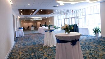 A spacious hall with a patterned carpet, round tables with white tablecloths and blue sashes, and large windows allowing natural light.